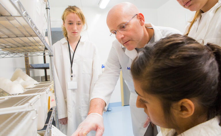 Instructor Abel Bult-It reads a lab paper held by a student, with two students looking on. The students and instructor are wearing white lab coats.